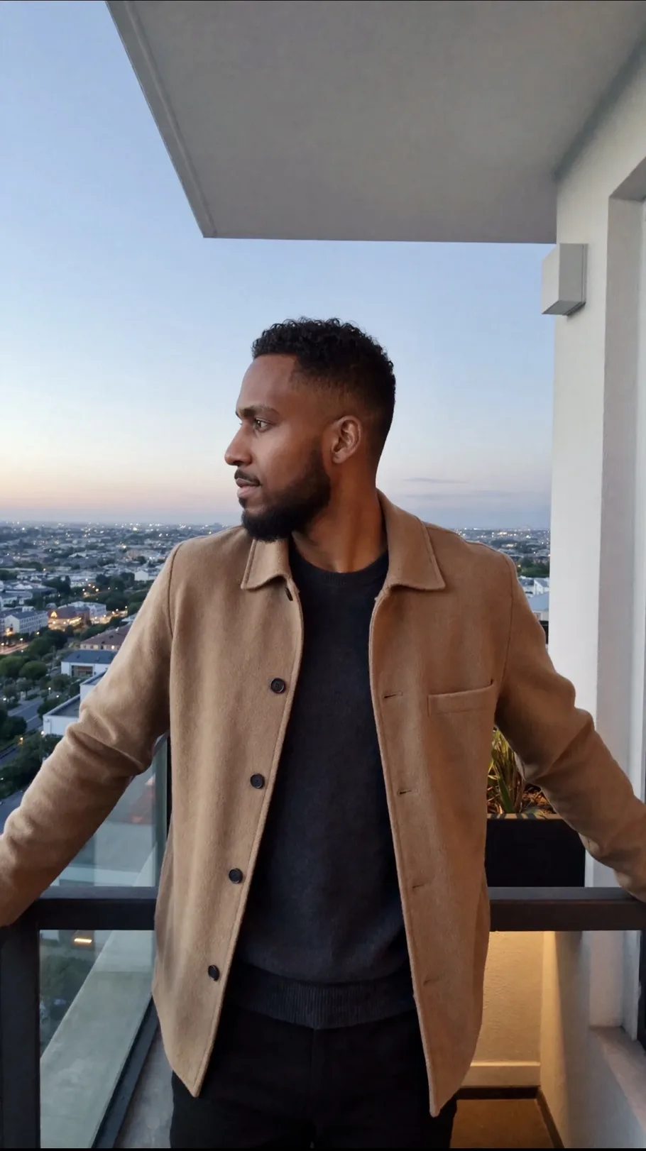 King The Kemist in a tan jacket on a high balcony at dusk, looking out over the city