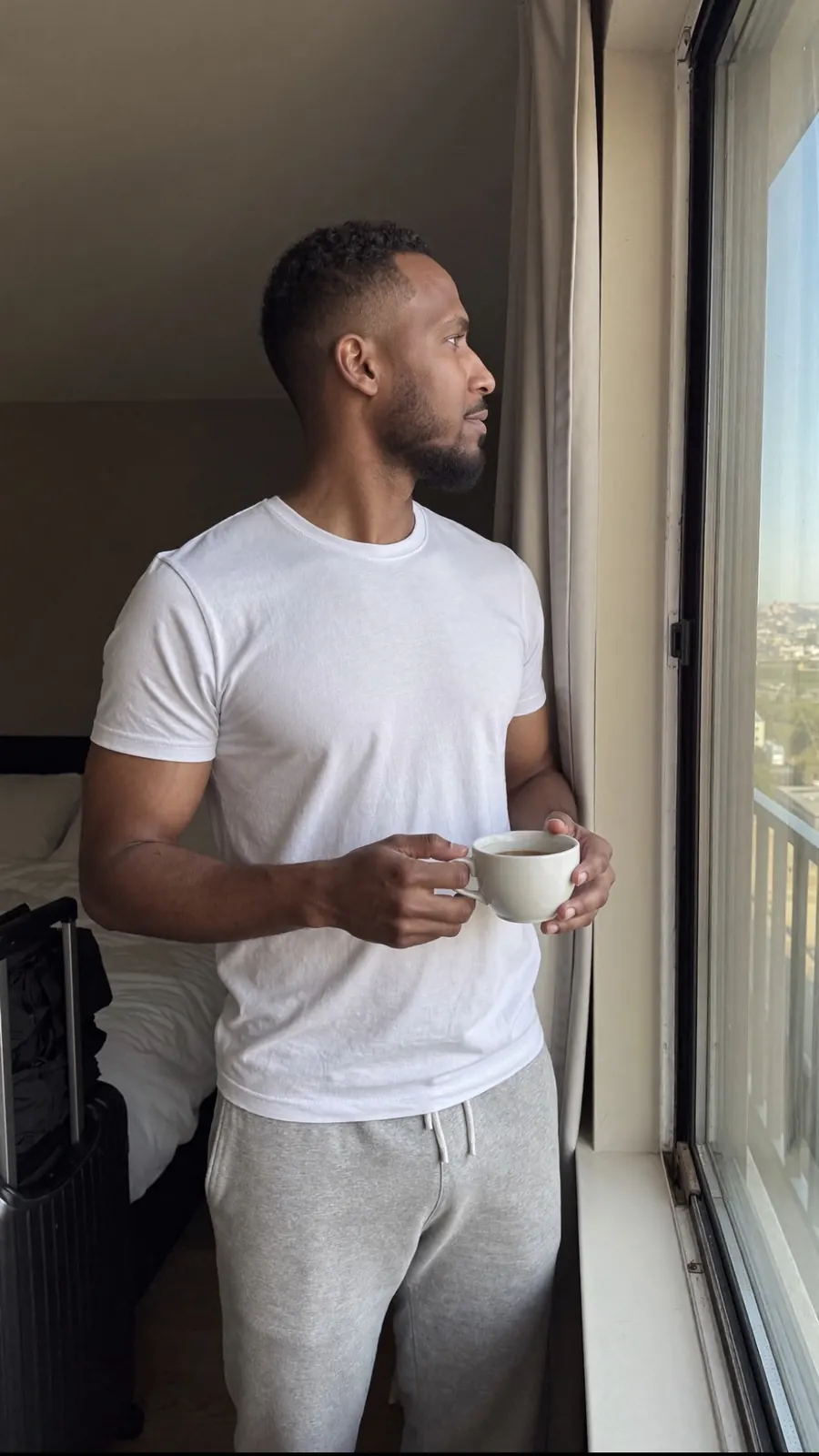King The Kemist in a white T-shirt holding a coffee cup and looking out a window