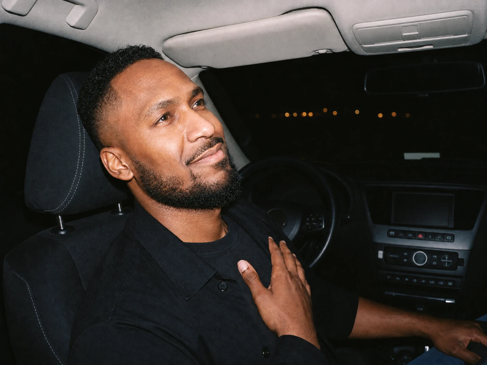 King The Kemist sits in the driver's seat of a parked car at night, looking upward with one hand resting on his chest