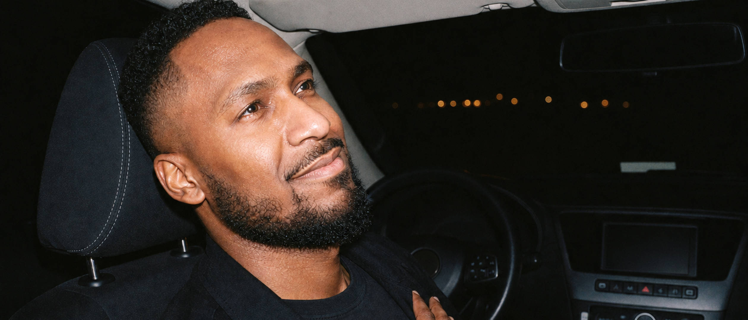 King The Kemist sits in the driver's seat of a parked car at night, looking upward with one hand resting on his chest