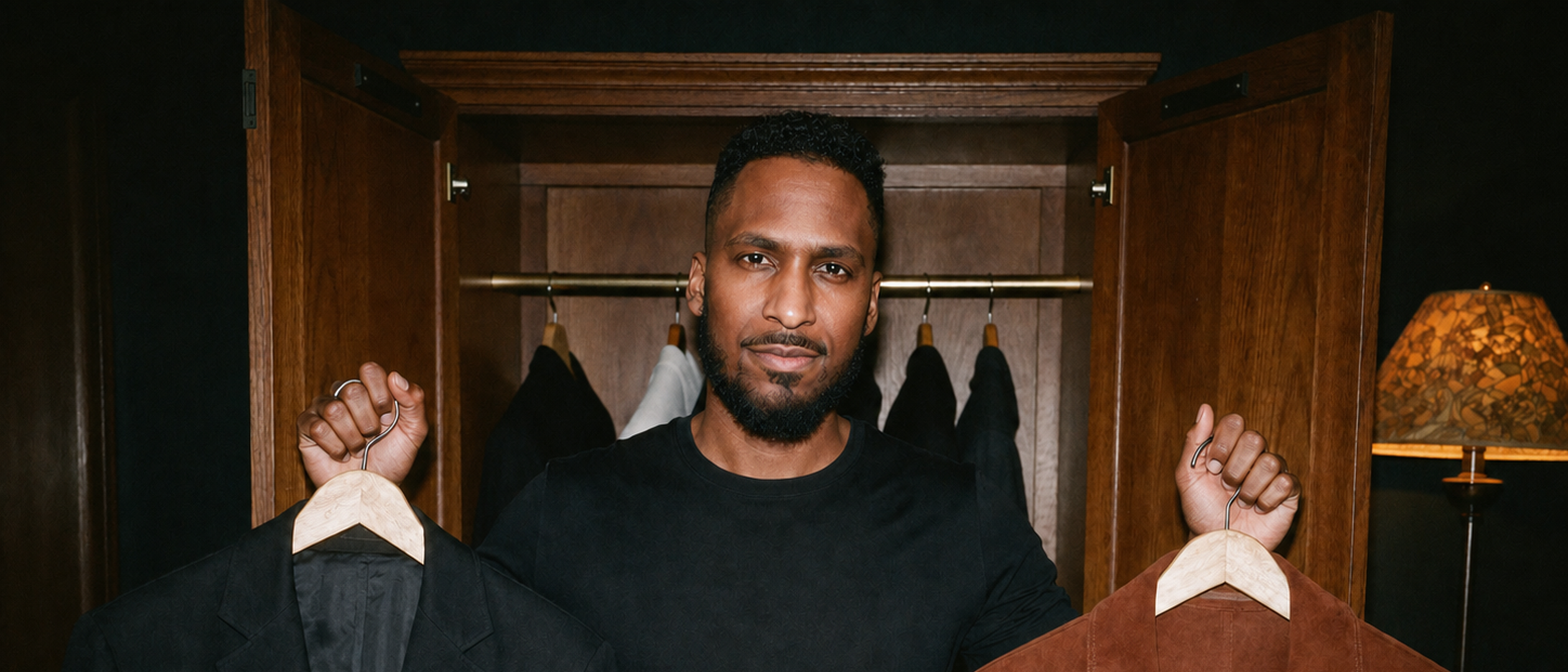 King The Kemist stands before an open wardrobe holding a black blazer and an amber suede jacket, weighing both options